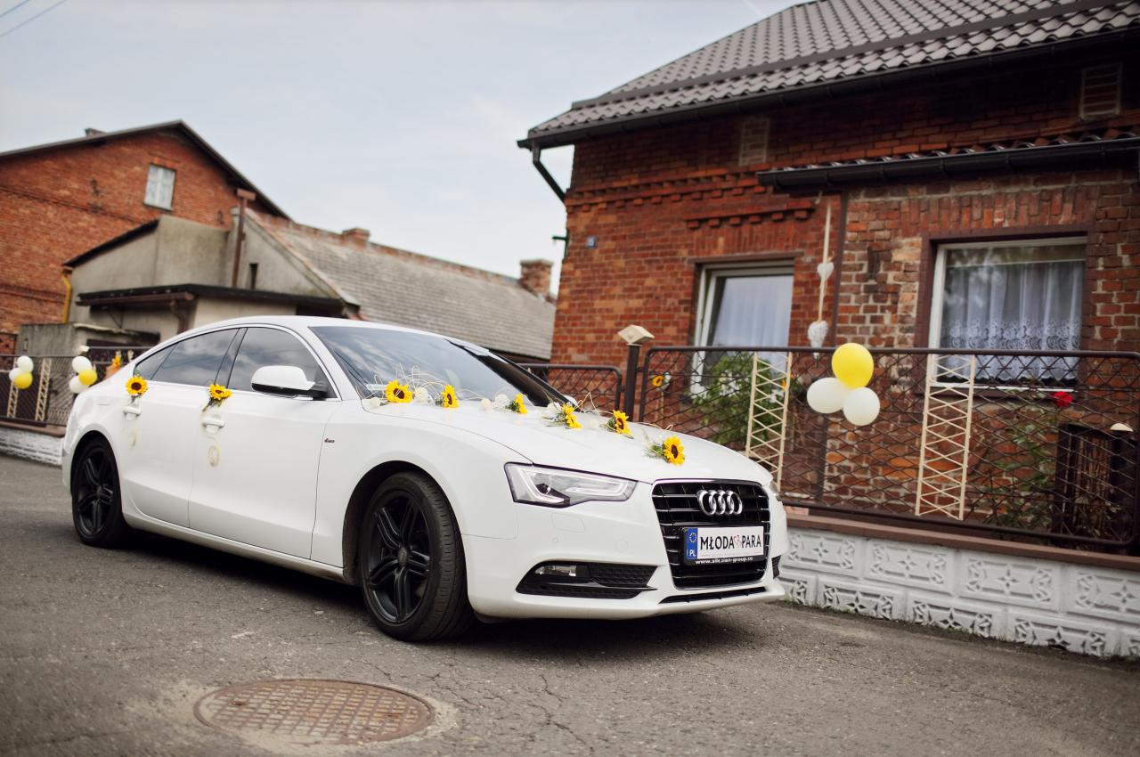 Białe Audi A5 udekorowane słonecznikami i balonami, tablica rejestracyjna z napisem 'Młoda Para', na tle ceglanego domu. Dekoracja ślubna samochodu.