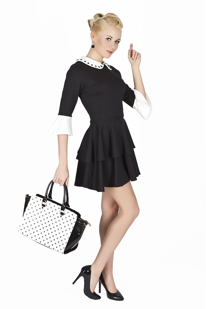 Elegancka kobieta w czarnej sukience z białym kołnierzykiem i mankietami, trzymająca torebkę w biało-czarne kropki, pozuje na białym tle.