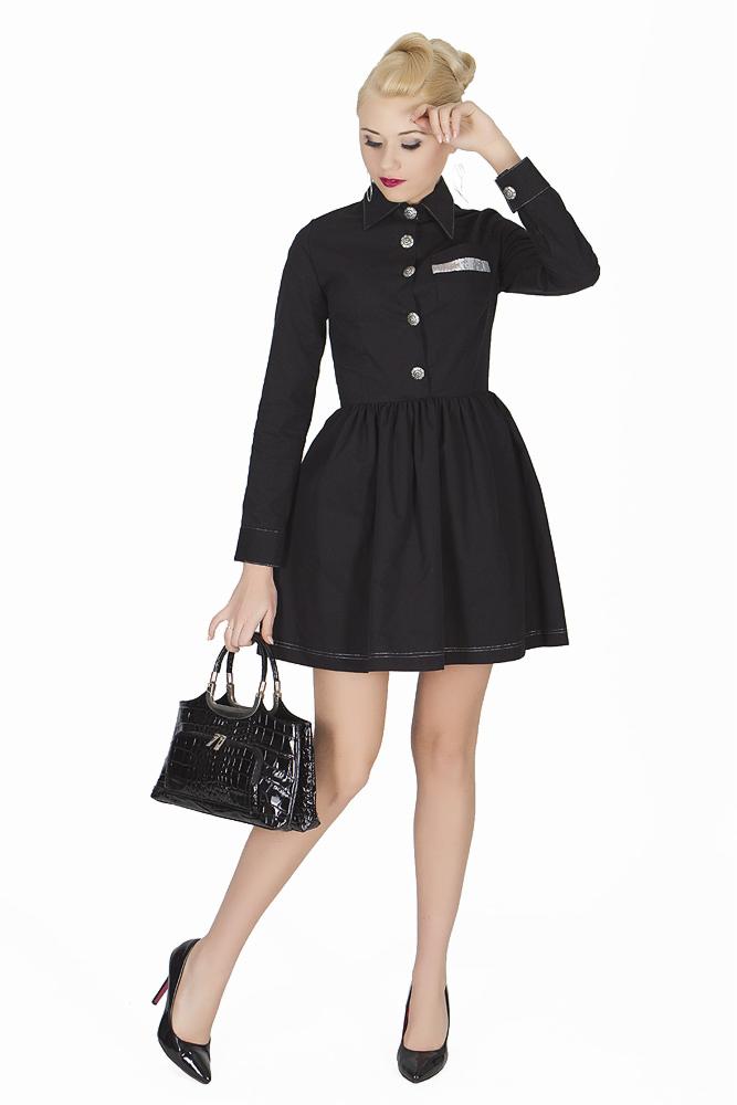 Kobieta w czarnej sukience z kołnierzykiem i torebką w stylu retro na białym tle, prezentująca stylizację z srebrnymi guzikami i wykończeniami.