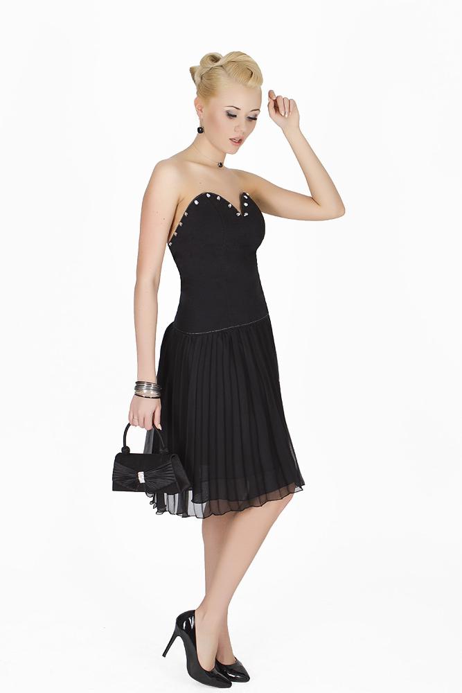 Elegancka blondynka w czarnej sukience bez ramiączek z plisowanym dołem, trzymająca małą torebkę, pozuje na białym tle. Całość uzupełniają czarne szpilki i biżuteria.