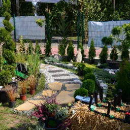 Aranżacja ogrodu pokazowa: ścieżka z kamieni, rośliny w donicach, system nawadniania na słomie. Różnorodność gatunków i materiałów w ogrodowej ekspozycji.