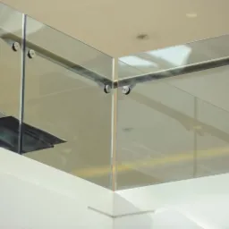 Szklana balustrada z metalowymi mocowaniami, widoczna z dołu, z elementami schodów w tle. Minimalistyczny design i jasne otoczenie.