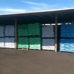 Magazyn zadaszony, palety z workami w kolorach: żółtym, białym, zielonym i niebieskim. Worki ułożone w rzędach, składowisko pod gołym niebem.