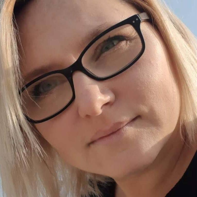 Portret kobiety z blond włosami i okularami w czarnych oprawkach, z delikatnym uśmiechem i niebieskimi oczami, w naturalnym oświetleniu.