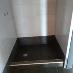 Wykończony brodzik z ciemnymi płytkami podłogowymi i jasnymi płytkami ściennymi, widoczny odpływ liniowy i częściowo przesuwne drzwi kabiny prysznicowej.