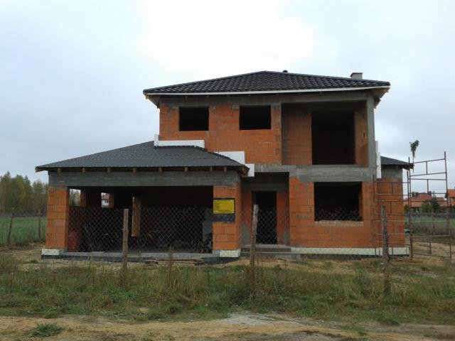 Dwukondygnacyjny dom z czerwonej cegły w trakcie budowy, z widocznym zadaszeniem garażu i czarnym dachem. Tło stanowią drzewa i pochmurne niebo.