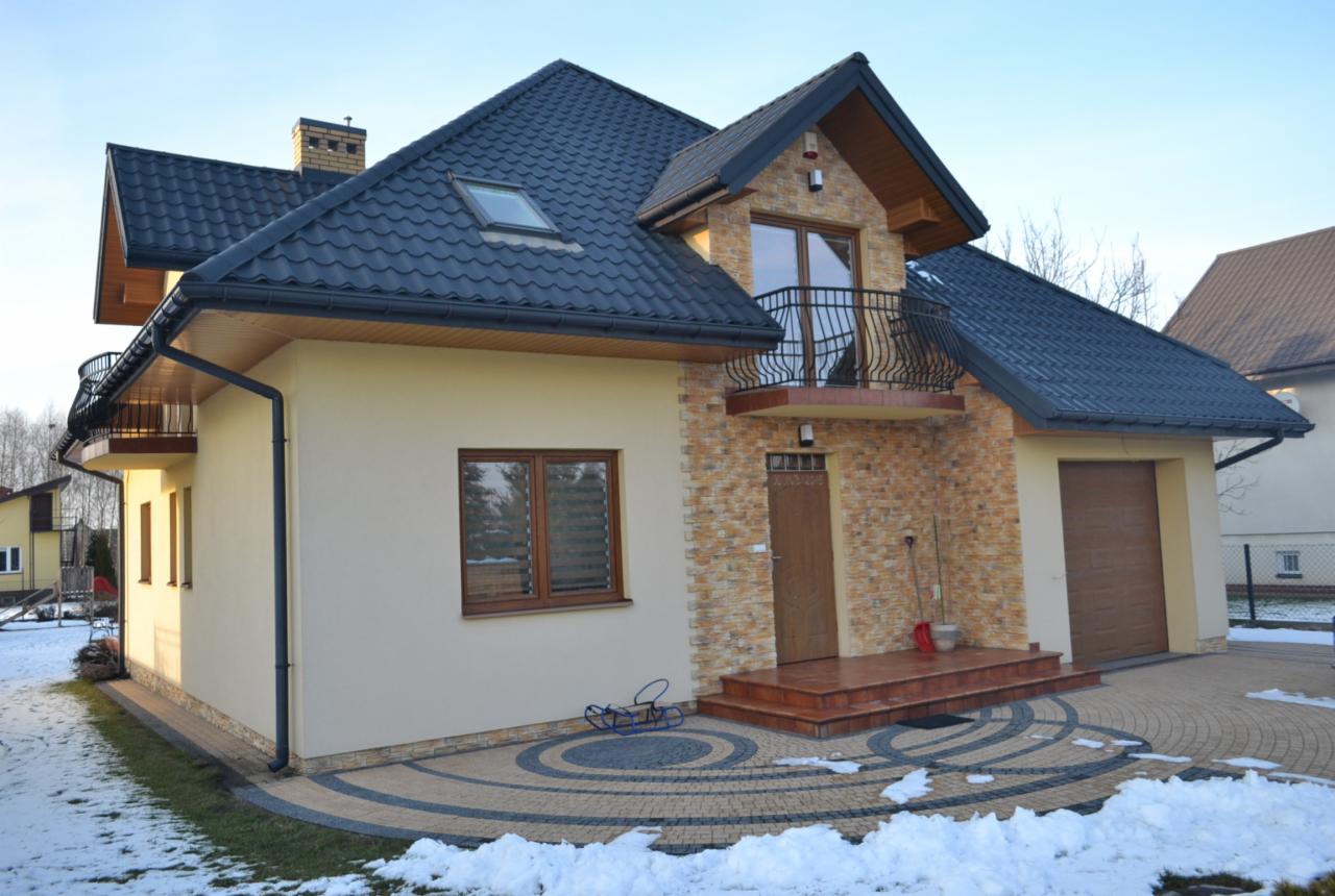 Dwukondygnacyjny dom z balkonem, wykończony beżowym tynkiem i kamieniem dekoracyjnym, z ciemną dachówką i kostką brukową na podjeździe. Zima, częściowo pokryty śniegiem.