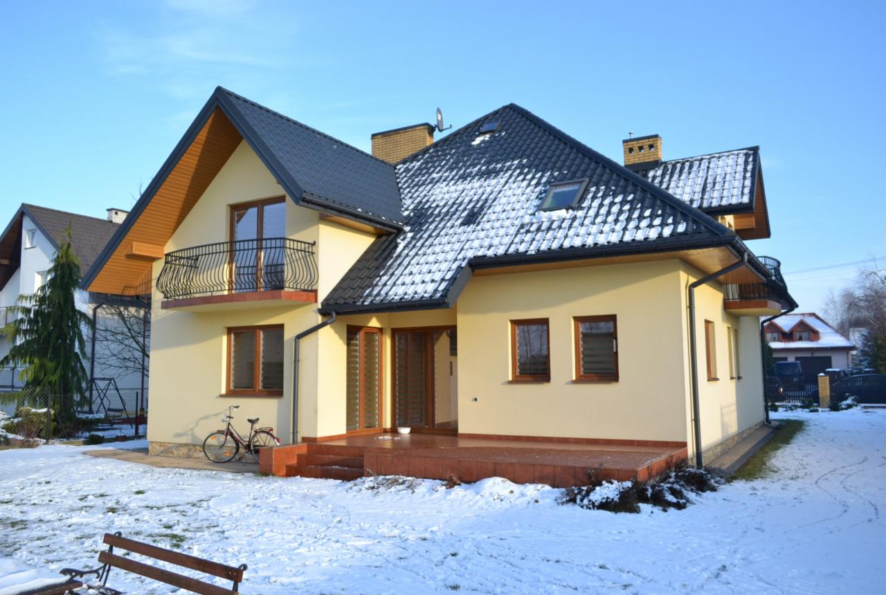 Otynkowany dom jednorodzinny z czarnym dachem pokrytym śniegiem, balkonem z kutą balustradą i tarasem z brązowych płytek. Na trawniku leży śnieg.