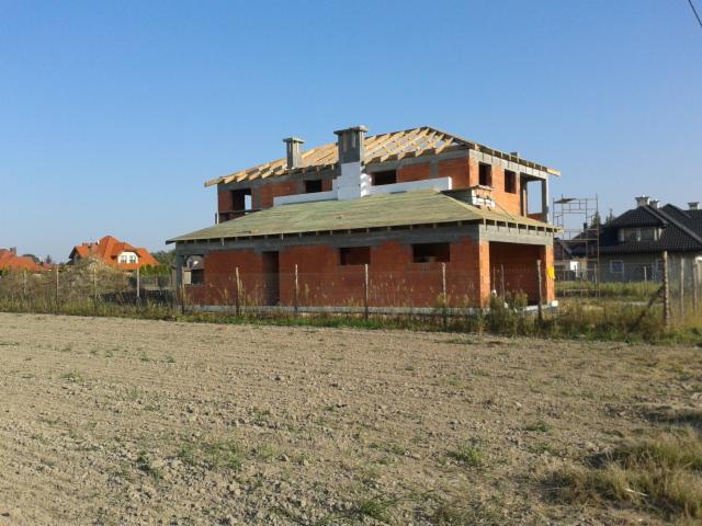 Surowy, dwukondygnacyjny dom z czerwonej cegły w trakcie budowy, z widoczną konstrukcją dachu, na tle błękitnego nieba i okolicznej zabudowy.