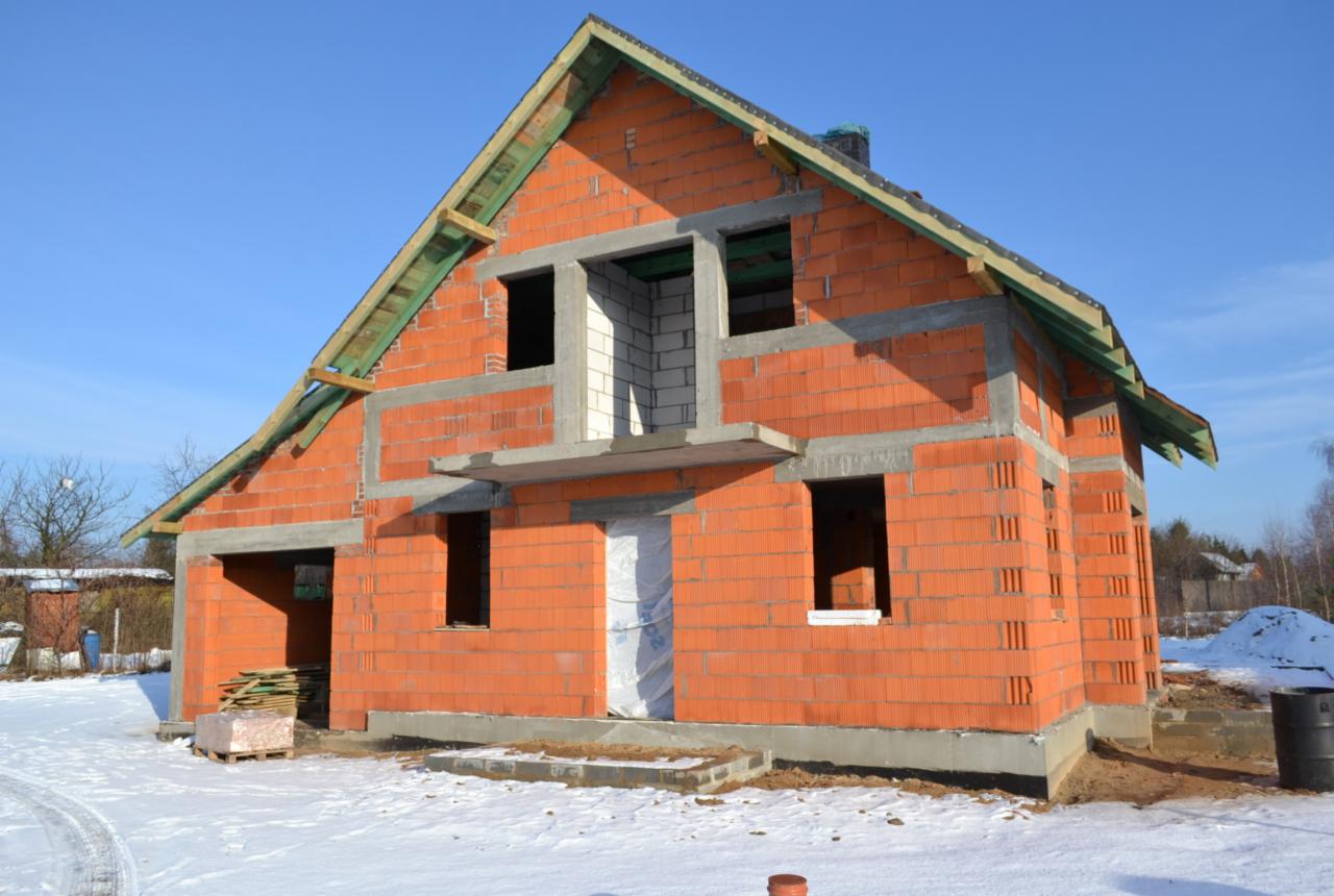 Niedokończony, dwukondygnacyjny dom murowany z czerwonej cegły porotherm, z widocznym zbrojeniem nad oknami i drzwiami, na tle zimowego krajobrazu i błękitnego nieba.