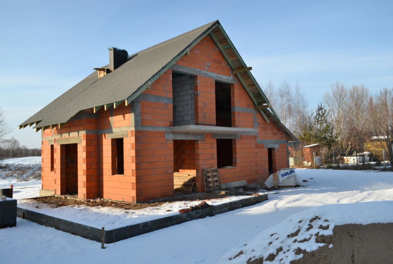 Surowy, dwukondygnacyjny dom z czerwonej cegły w trakcie budowy, z widocznymi otworami okiennymi i drzwiowymi, częściowo pokryty dachem, zimowy krajobraz w tle.