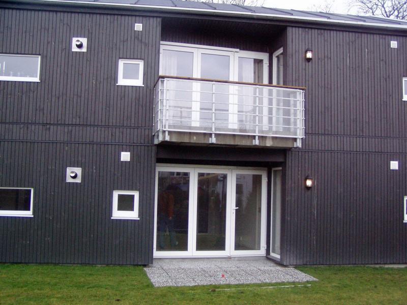 Elewacja budynku z ciemnego drewna z balkonem i dużymi, białymi oknami. Widoczne detale architektoniczne i oświetlenie zewnętrzne.