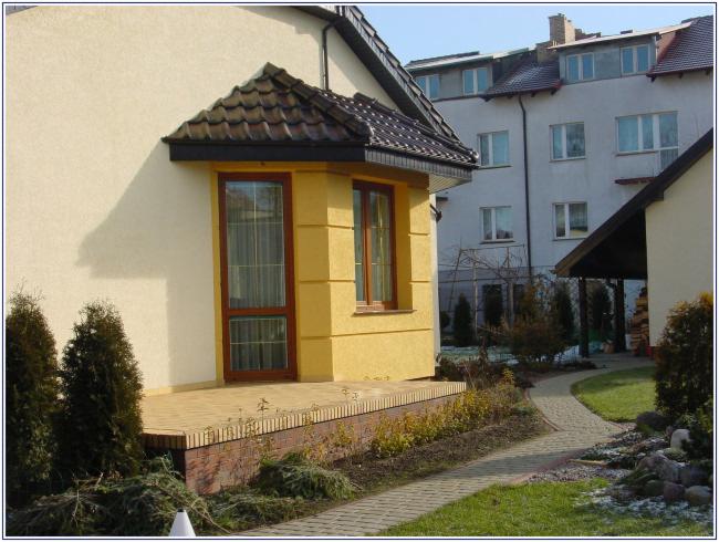 Żółta dobudówka z oknami z brązowymi ramami i zasłonami, widok z zewnątrz, na tle domu i ścieżki z kostki.