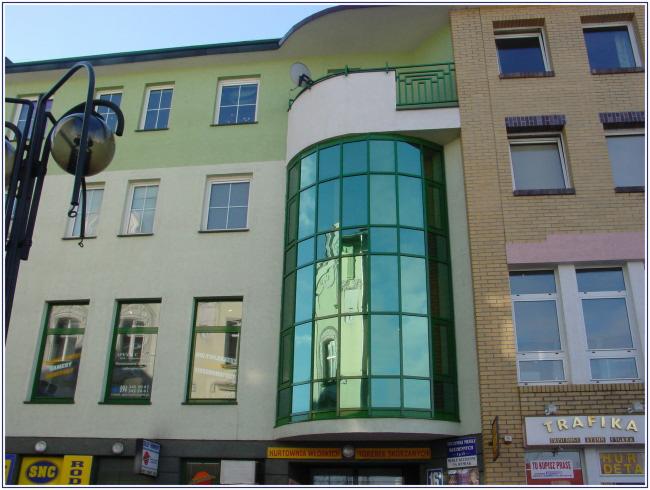 Elewacja budynku z różnymi typami okien: standardowe, okrągłe, oraz z zielonego szkła. Widoczny fragment ulicy i szyldy sklepów na dole budynku.