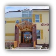 Żółty budynek z ciemnoczerwoną cegłą wokół wejścia i nowoczesnymi oknami. Nad wejściem drewniana konstrukcja daszku. W tle błękitne niebo.