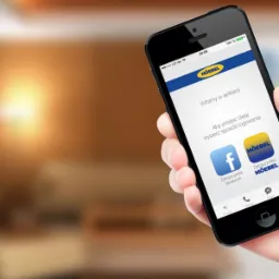 Aplikacja mobilna na smartfonie prezentująca interfejs logowania do platformy MÖEBEL, trzymana w dłoni na tle rozmytego wnętrza.