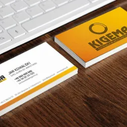 Dwie wizytówki firmy Kigema leżą obok białej klawiatury na drewnianym biurku. Jedna wizytówka jest w kolorze żółtym, druga biała z pomarańczowym akcentem.