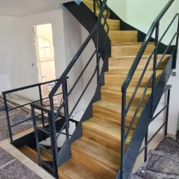 Nowoczesne schody z drewnianymi stopniami i metalową, czarną konstrukcją w surowym wnętrzu, widoczne zabezpieczenia podłogi podczas prac wykończeniowych.