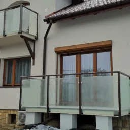 Nowoczesny dom z balkonami z matowego szkła i ciemnymi ramami, widoczne elementy wykończenia z drewnopodobnych paneli oraz kamienia elewacyjnego, na pierwszym planie lekko oszronione krzewy.