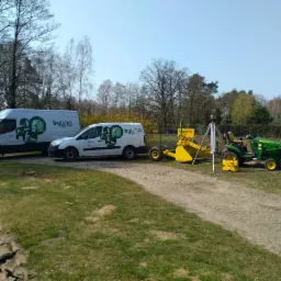 Dwa białe samochody dostawcze z logo firmy, żółta niwelarka laserowa oraz zielony traktor z łyżką na tle drzew i trawy.