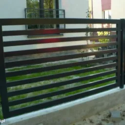 Nowoczesne, poziome ogrodzenie panelowe w kolorze antracytowym, z widocznym fragmentem budynku w tle. Minimalistyczny design i precyzyjne wykonanie.