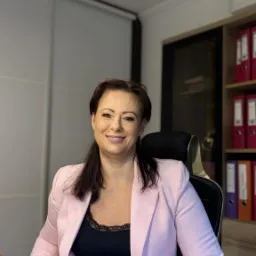 Uśmiechnięta kobieta w różowej marynarce siedzi przy biurku w biurze, za nią regał z segregatorami i przesuwane drzwi szafy.