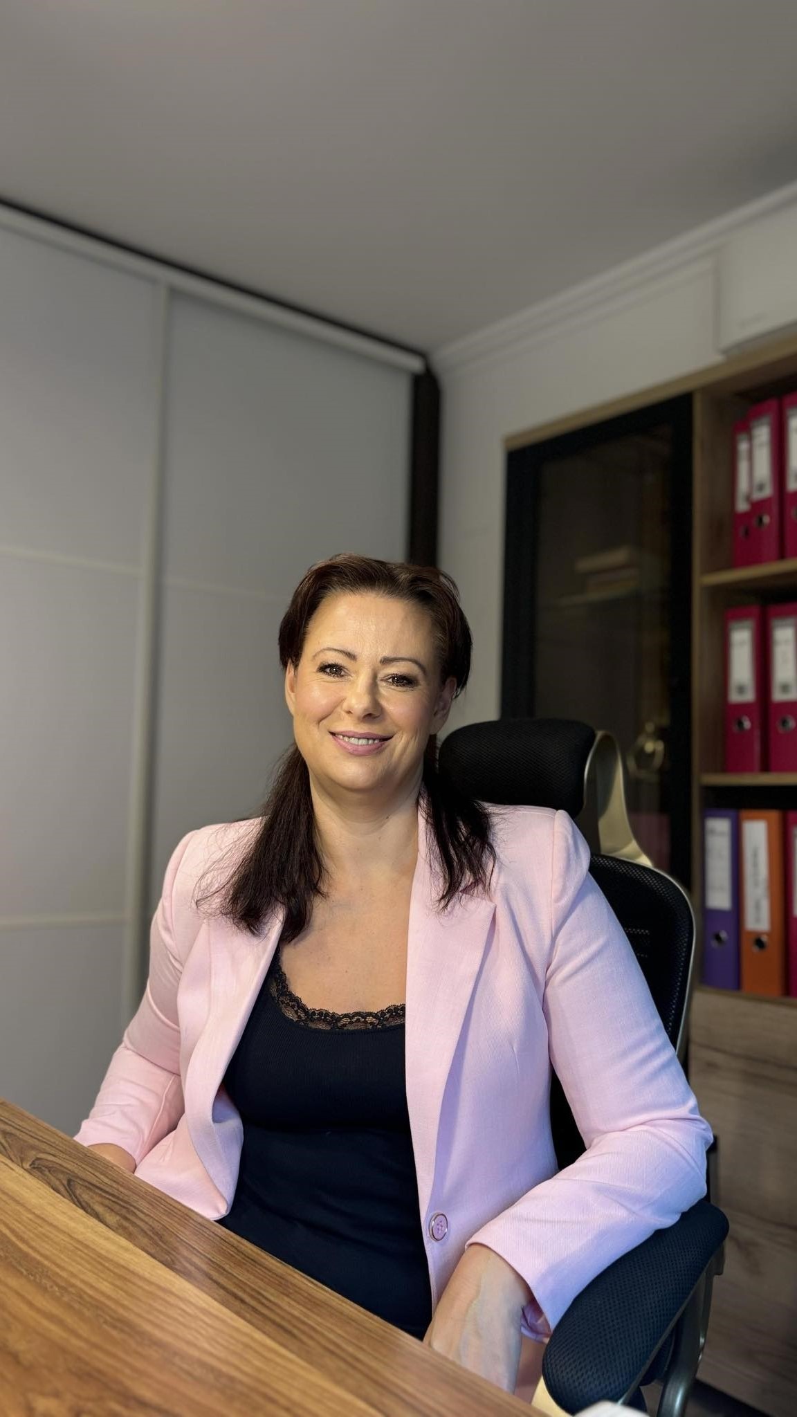 Uśmiechnięta kobieta w różowej marynarce siedzi przy biurku w biurze, za nią regał z segregatorami i przesuwane drzwi szafy.