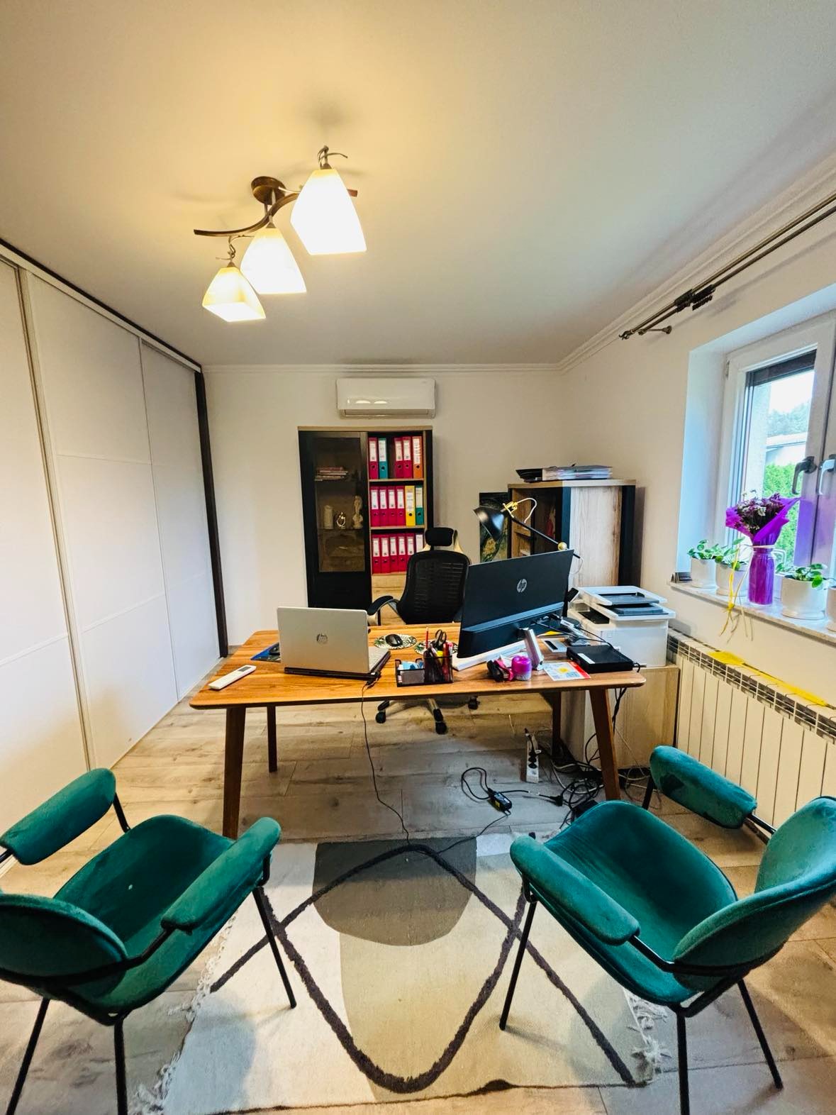 Biuro z drewnianym biurkiem, dwoma zielonymi fotelami, komputerami, regałem z segregatorami i kwiatami w doniczkach na parapecie