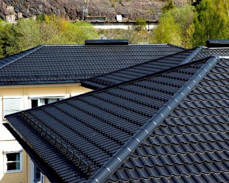 Czarny, metalowy dach dwuspadowy z rynnami i zabezpieczeniami śniegowymi na tle budynku z oknami i zieleni, widok z góry, oświetlenie dzienne.