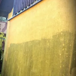 Żółta elewacja budynku z widocznymi zabrudzeniami i mchem, balkon z drewnianą balustradą w tle. Ukazany fragment ściany przed czyszczeniem.