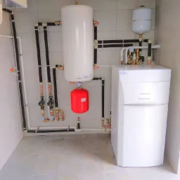 Biała pompa ciepła De Dietrich z podłączonymi rurami, zbiornikiem buforowym i naczyniem przeponowym w pomieszczeniu z szarymi płytkami.