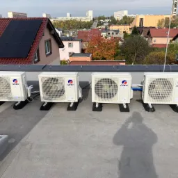 Cztery jednostki zewnętrzne klimatyzacji marki Gree zamontowane na płaskim dachu, z widocznymi niebieskimi przewodami i narzędziami, w tle zabudowa miejska i domy z panelami słonecznymi.