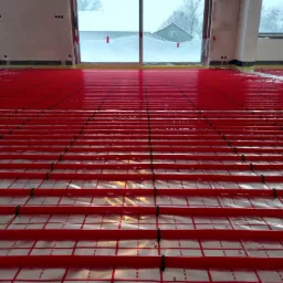 Czerwone rury ogrzewania podłogowego rozłożone na folii z nadrukowaną siatką, widok z niskiej perspektywy w kierunku dużego okna z widokiem na zaśnieżony krajobraz.