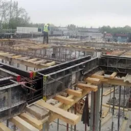 Szeroki kadr na budowę stropu z widocznymi szalunkami, drewnianymi podporami i zbrojeniami, z dwoma pracownikami w odblaskowych kamizelkach i żółtych kaskach na górze konstrukcji, w tle zabudowa...