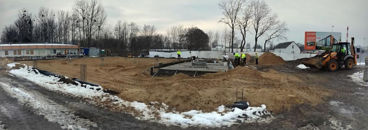 Wykop pod fundamenty budynku usługowego w trakcie prac ziemnych; widoczny szalunek, zbrojenie, pracownicy w kamizelkach odblaskowych oraz koparko-ładowarka.