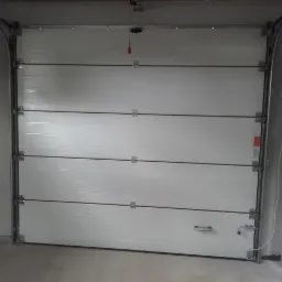 Biała segmentowa brama garażowa z poziomymi przetłoczeniami, zamontowana w surowym garażu z betonowymi ścianami. Widoczne elementy mechanizmu otwierania.