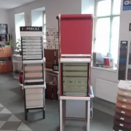 Ekspozycja dwóch stojaków z roletami okiennymi w sklepie, z widocznymi próbkami kolorów i wzorów, na tle jasnego wnętrza z oknami i biurkiem.