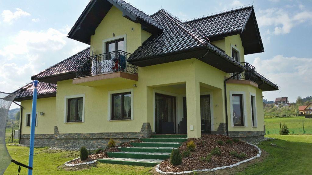 Żółty dom dwupiętrowy z ciemnym dachem, balkonami z kutymi balustradami i kamienną elewacją na zielonym trawniku. Widok na okolicę w tle.