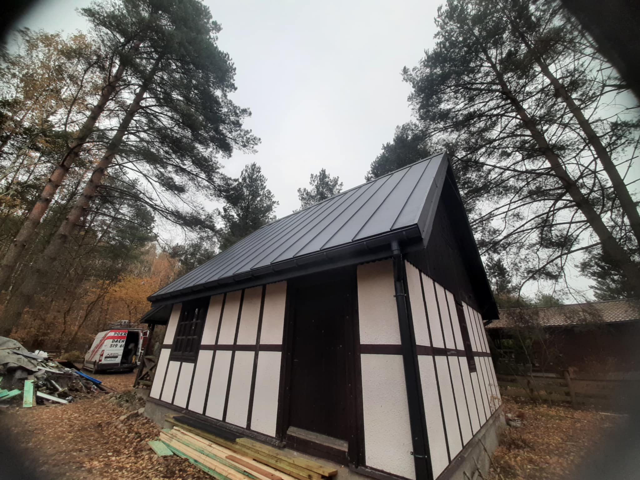 Mały budynek z ciemnym dachem z blachy na tle drzew, z widocznym samochodem dostawczym i resztkami materiałów budowlanych obok.