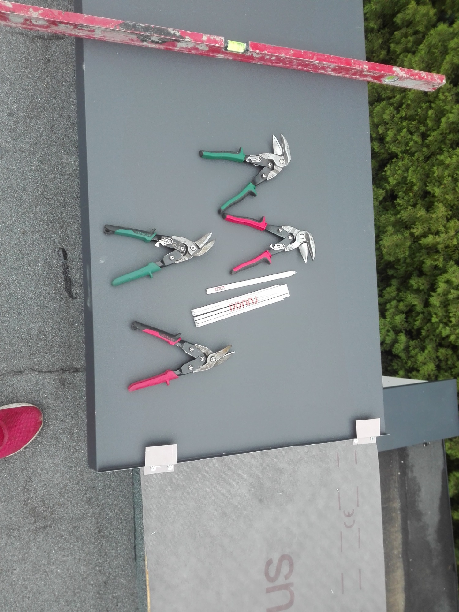 Szary panel dachowy z narzędziami dekarskimi: trzema parami nożyc do blachy Stanley z zielonymi i różowymi uchwytami, poziomicą, miarkami zwijanymi Toughbuilt, oraz fragmentem membrany dachowej...