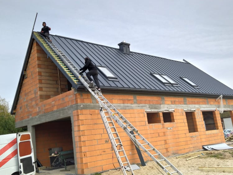 Dwóch dekarzy na drabinach montuje czarne pokrycie dachowe z oknami dachowymi na nowo budowanym domu z czerwonej cegły.