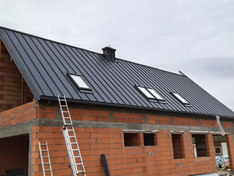 Nowo położony, grafitowy dach z trzema oknami dachowymi na budynku w stanie surowym otwartym, z cegły, z dwiema rozstawionymi drabinami opartymi o ścianę.
