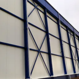 Boczny widok hali magazynowej z białymi panelami i niebieską konstrukcją stalową, widoczne elementy mocujące i niewielki stos blachy na ziemi.