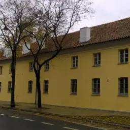 Długi, dwukondygnacyjny budynek o żółtej elewacji z regularnie rozmieszczonymi oknami i brązowym dachem z białymi kominami, widziany z perspektywy ulicy, z drzewami na pierwszym planie.