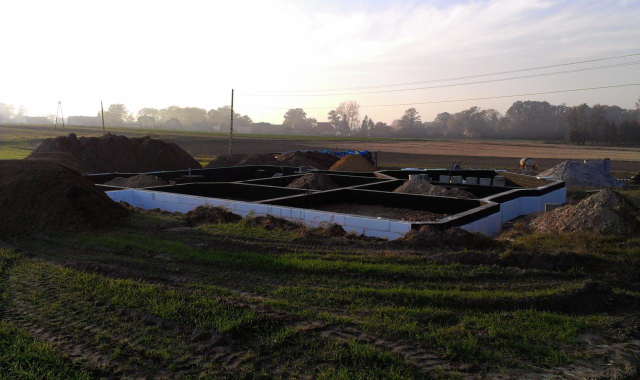 Przygotowanie fundamentów pod budowę domu jednorodzinnego: wylane ławy fundamentowe z izolacją termiczną, częściowo zasypane ziemią, widoczne stosy ziemi w otoczeniu, pole w tle.