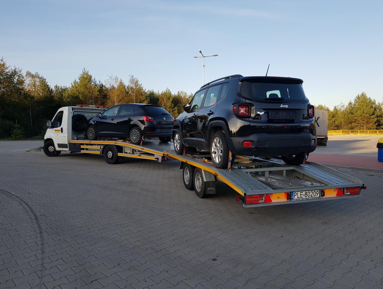 Dwa czarne samochody, Jeep i Seat, zabezpieczone na żółtej lawecie transportowej z widoczną tablicą rejestracyjną, na tle parkingu i drzew