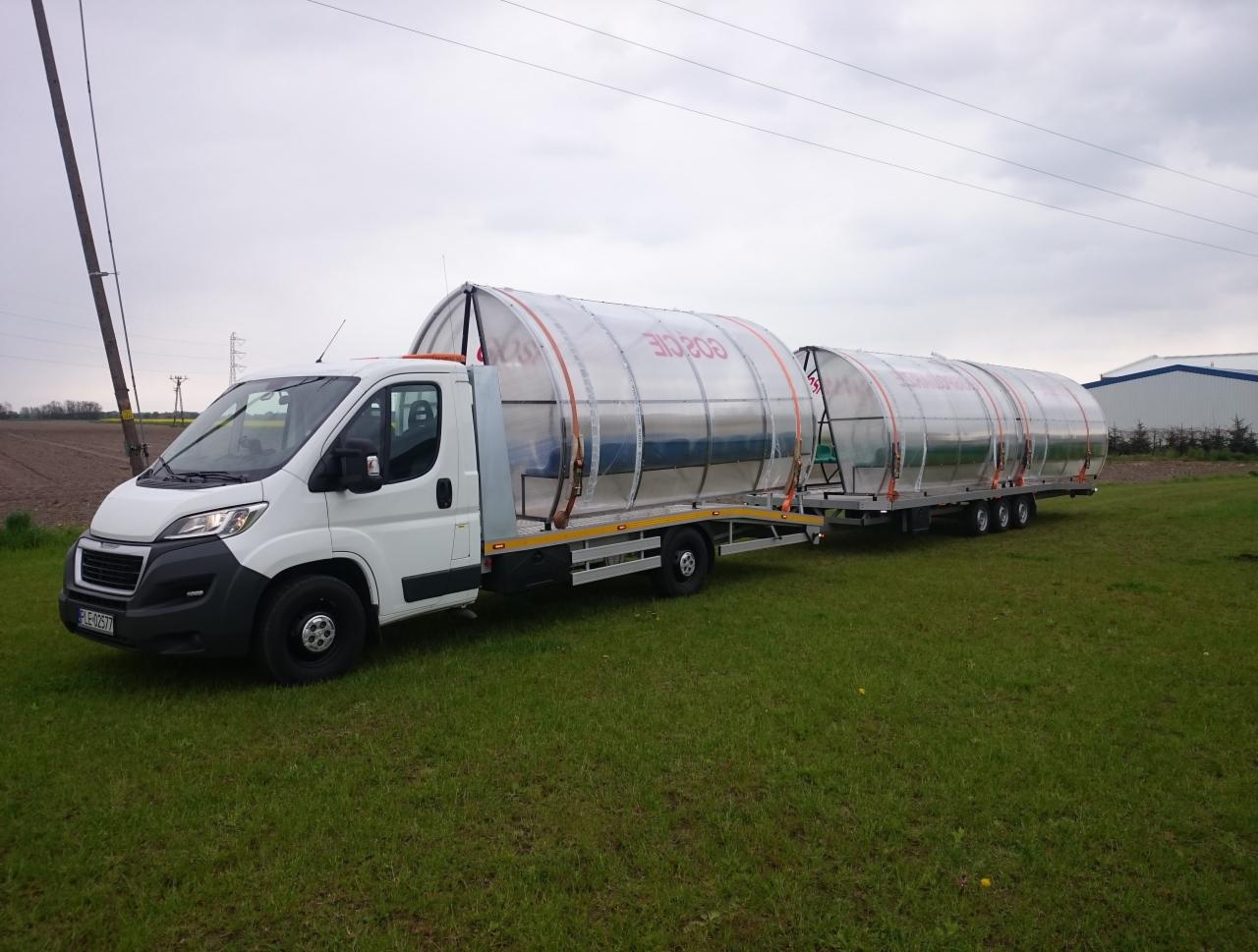 Biała laweta z długą przyczepą przewozi konstrukcję tunelu foliowego z napisem. Widok z boku na trawiastym terenie.