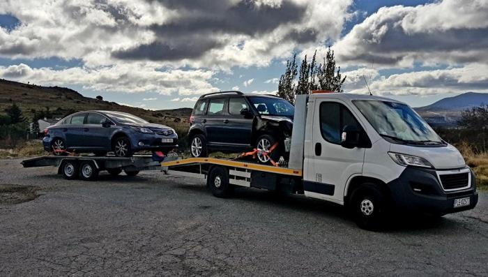 Biała laweta z dwoma samochodami: uszkodzonym SUV-em zabezpieczonym pasami i sedanem na przyczepie, na tle pagórkowatego krajobrazu i pochmurnego nieba.