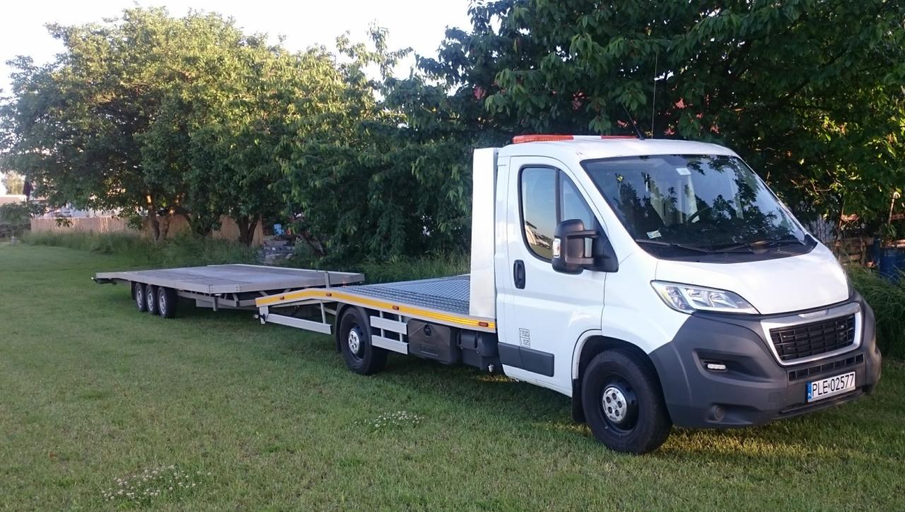 Biała laweta z niskopodwoziową przyczepą na trawiastym terenie, widoczne drzewa w tle.