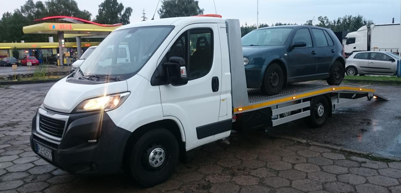 Biała laweta Peugeot z niebieskim samochodem osobowym na platformie, zaparkowana na parkingu stacji benzynowej Lotos w pochmurny dzień.